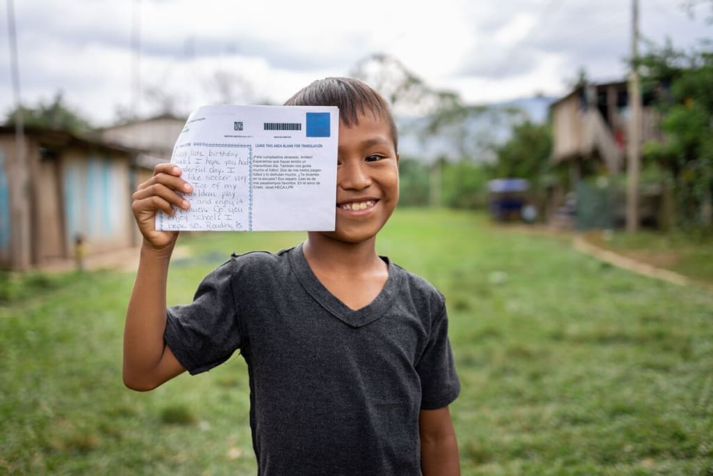 Anllelo is wearing a black shirt. He is standing in a grass field in his neighborhood and is holding up a letter.